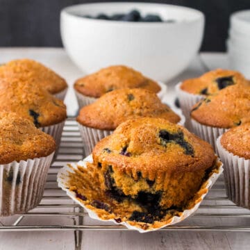 A batch of blueberry muffins sits on a cooling rack, with one muffin unwrapped to reveal the blueberries inside. A bowl of blueberries is in the background.