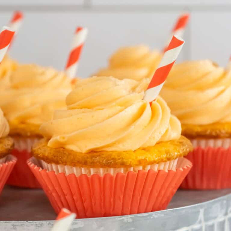 Orange soda cupcakes with orange frosting in orange wrappers, each topped with a red and white striped straw, displayed on a tray.