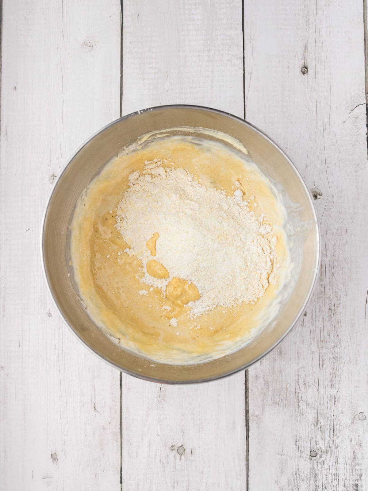 A metal mixing bowl with flour partially mixed into a yellow batter, sitting on a white wooden surface.