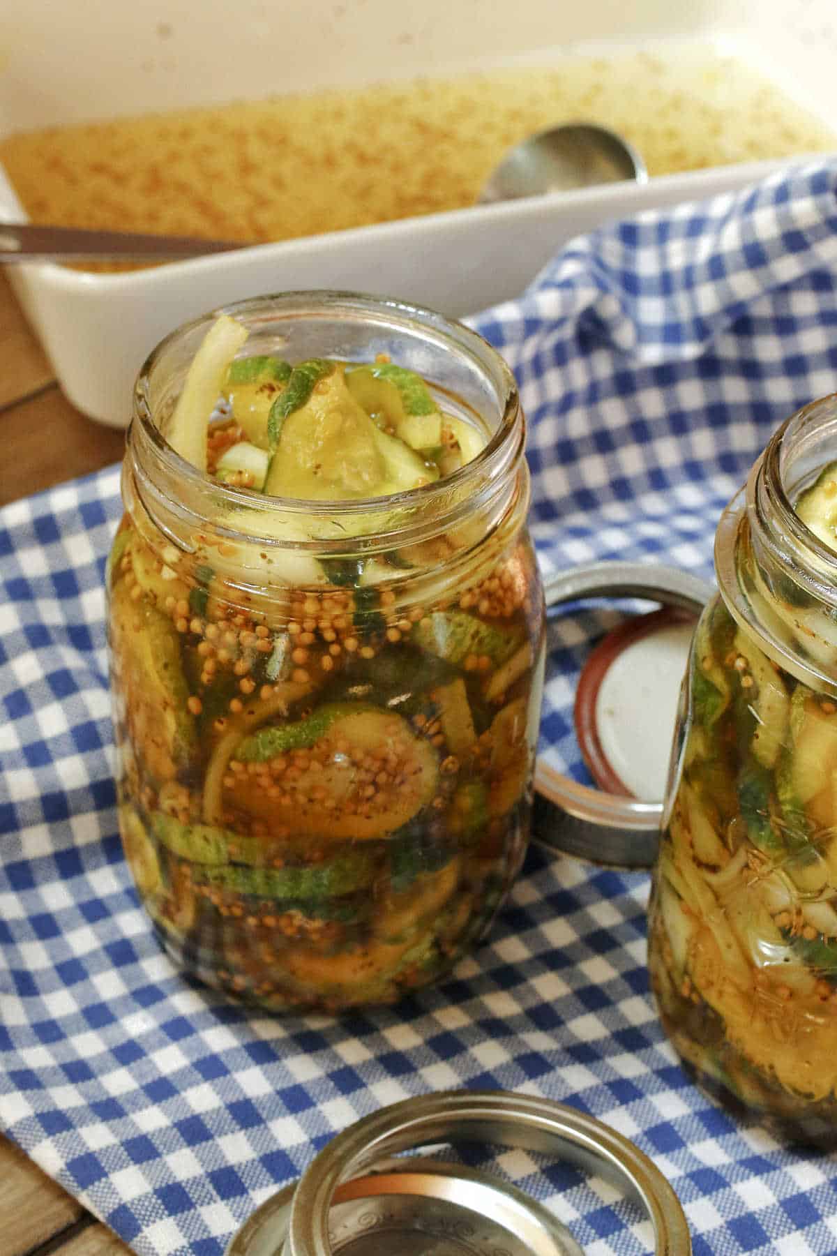 Two glass jars filled with pickled cucumbers and spices sit on a blue and white checkered cloth, with a dish of brine and a metal spoon in the background.