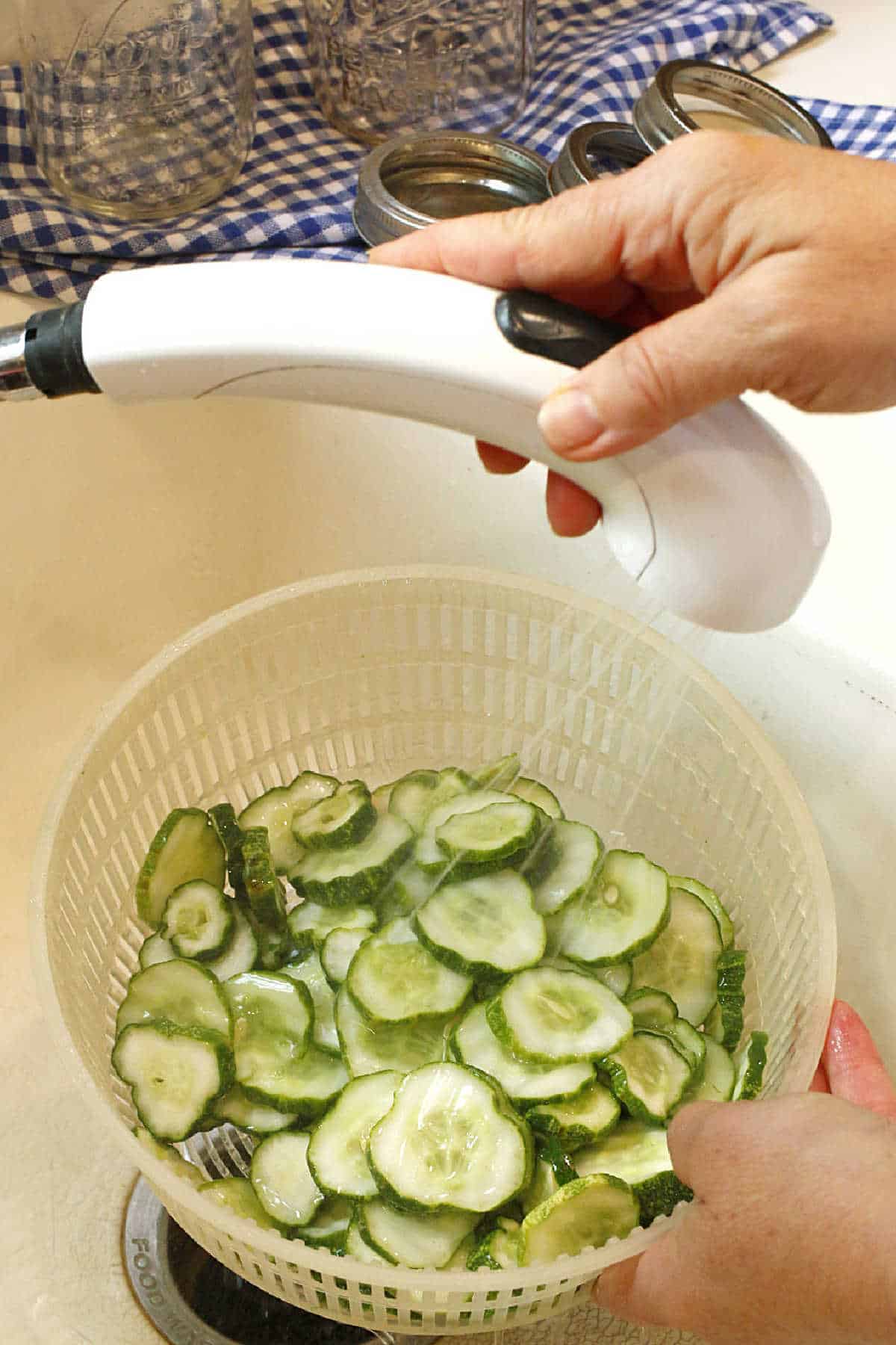 Sliced cucumbers being rinsed under running water in a white plastic colander, with canning jars and lids visible in the background.