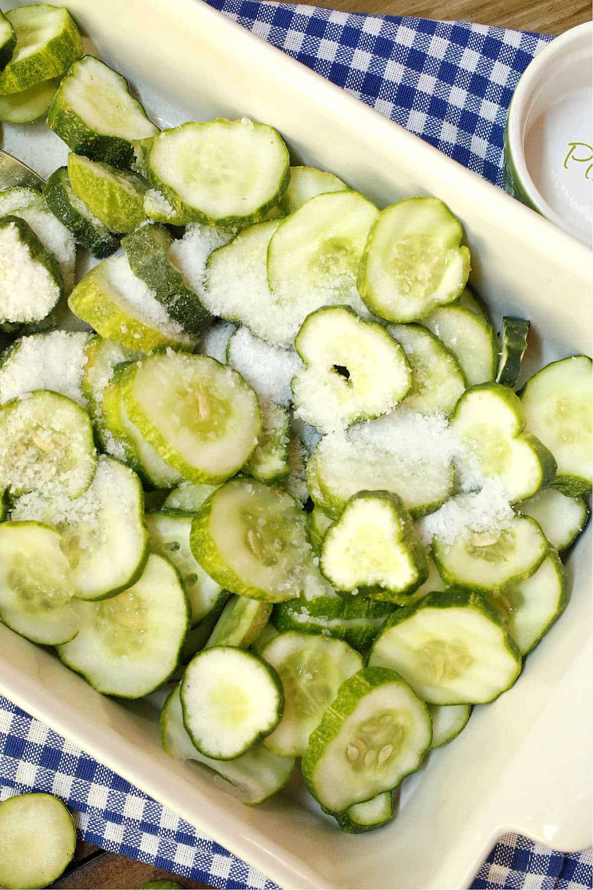 Sliced cucumbers sprinkled with salt in a rectangular dish, placed on a blue and white checkered cloth.