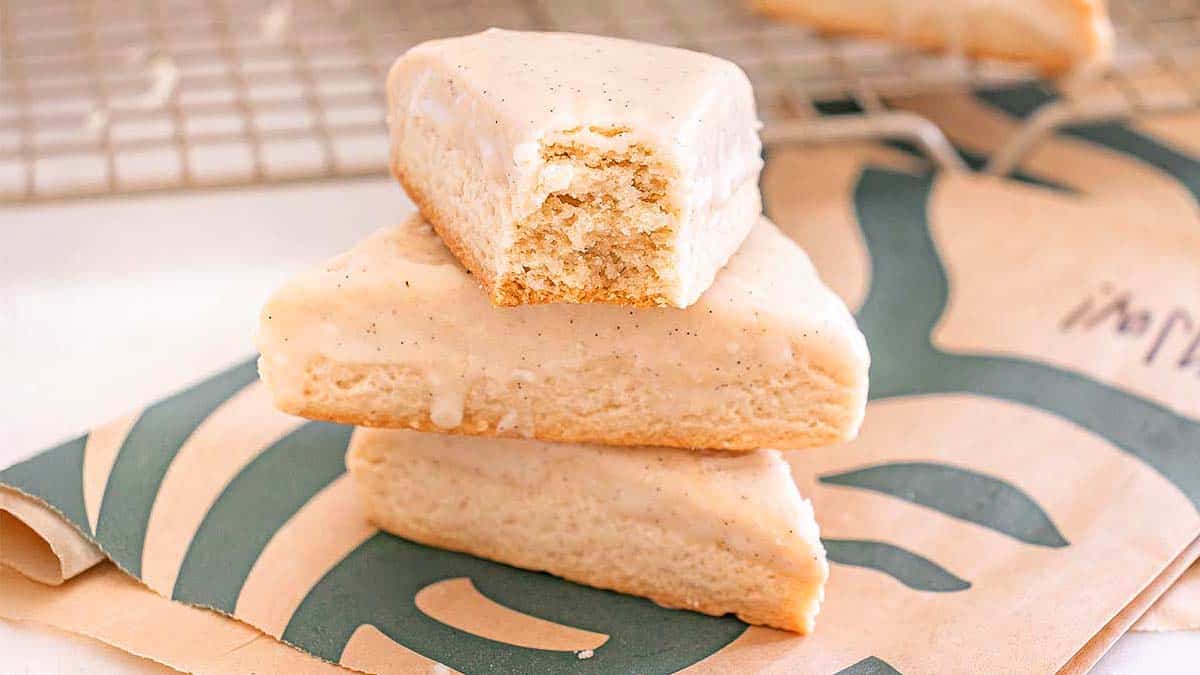Three vanilla iced scones are stacked on a Starbucks-branded bag; the top scone has a bite taken out of it.