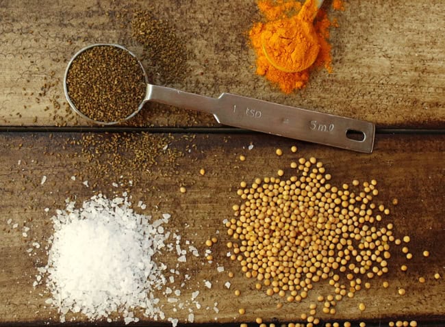 A metal measuring spoon with brown seeds, ground turmeric, coarse salt, and whole yellow mustard seeds on a wooden surface.