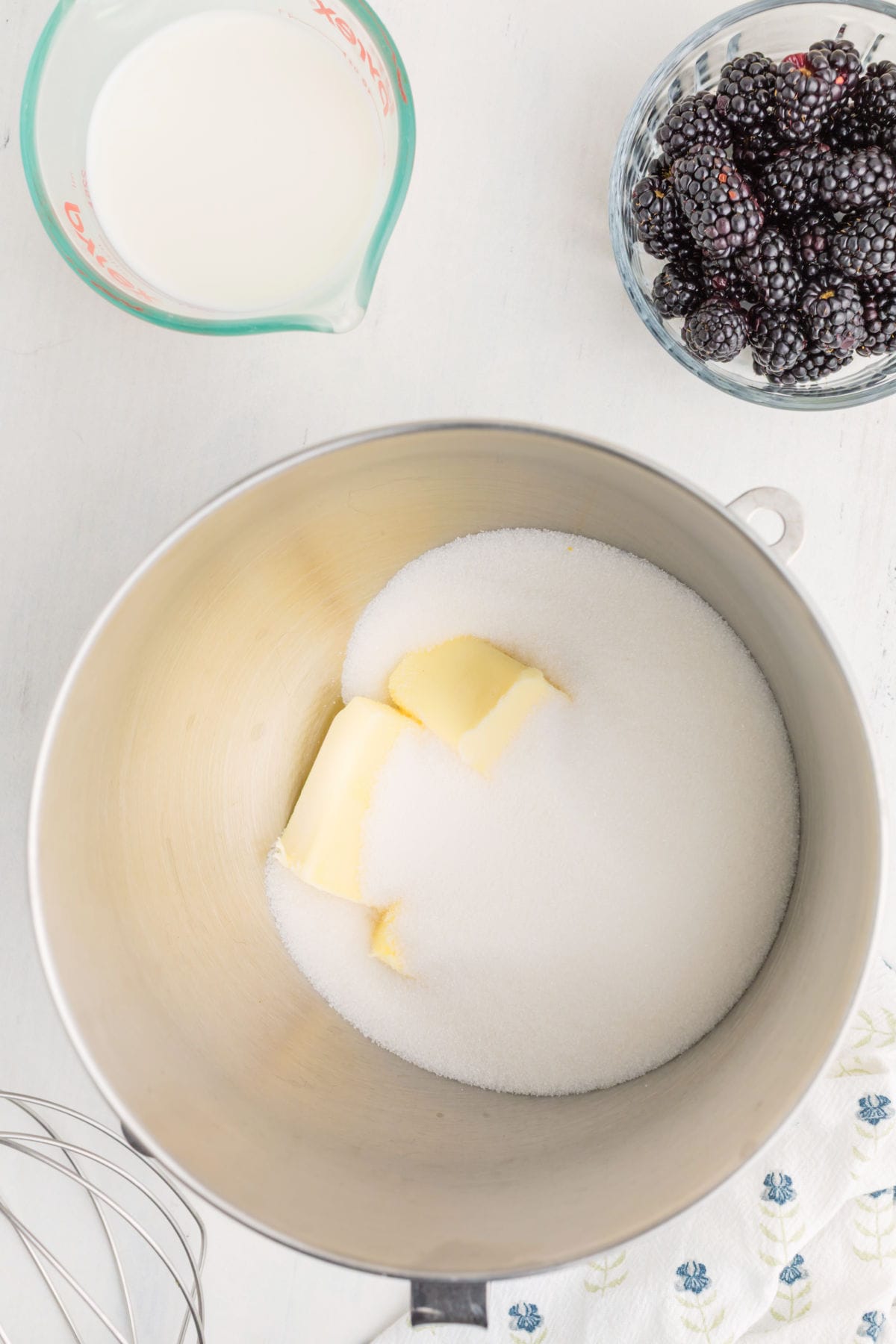 A mixing bowl with butter and sugar, a glass measuring cup of milk, a bowl of blackberries, and a whisk on a white surface.