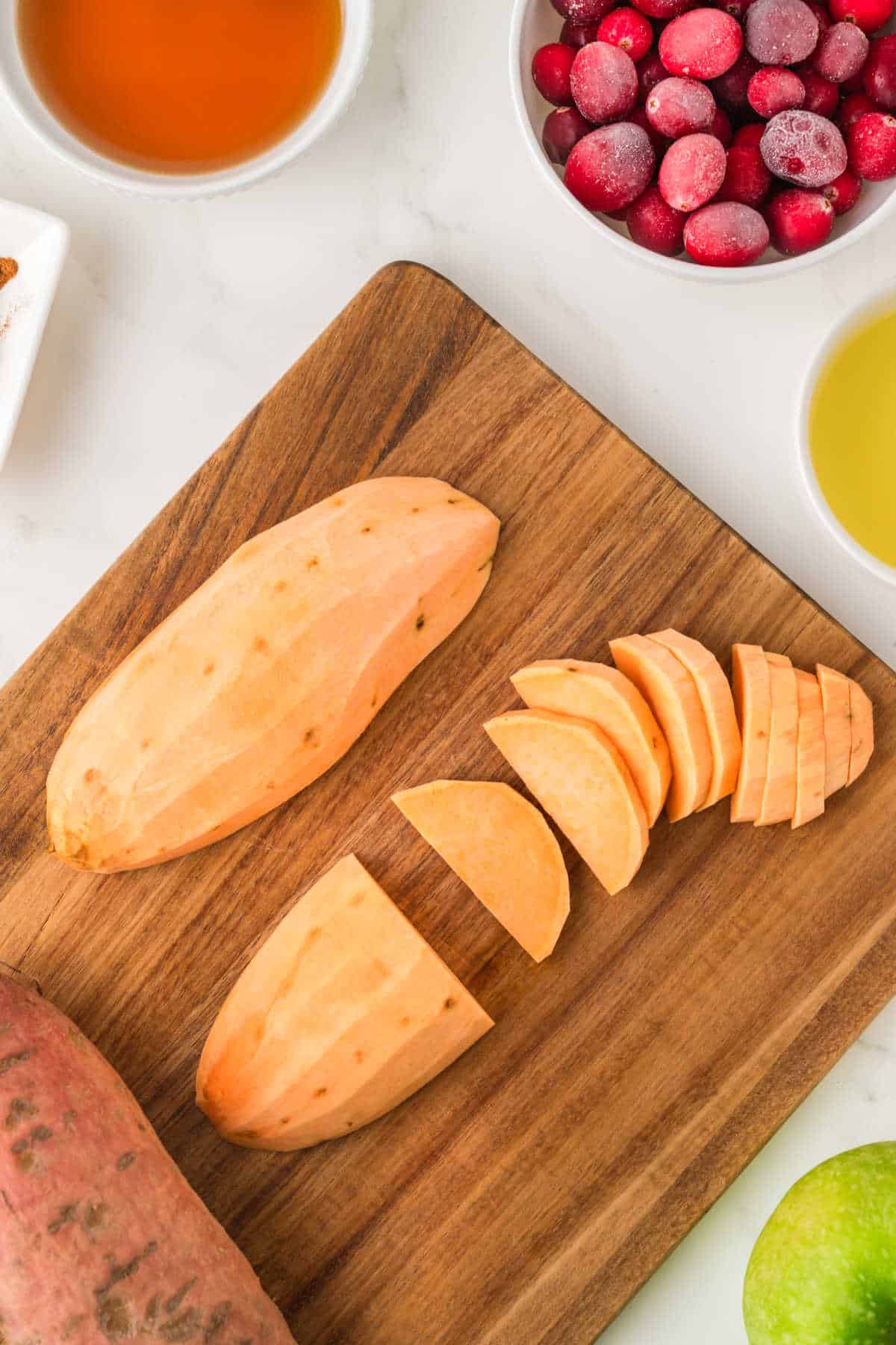 Peeled and sliced sweet potato on a wooden cutting board with bowls of cranberries, oil, and syrup nearby.