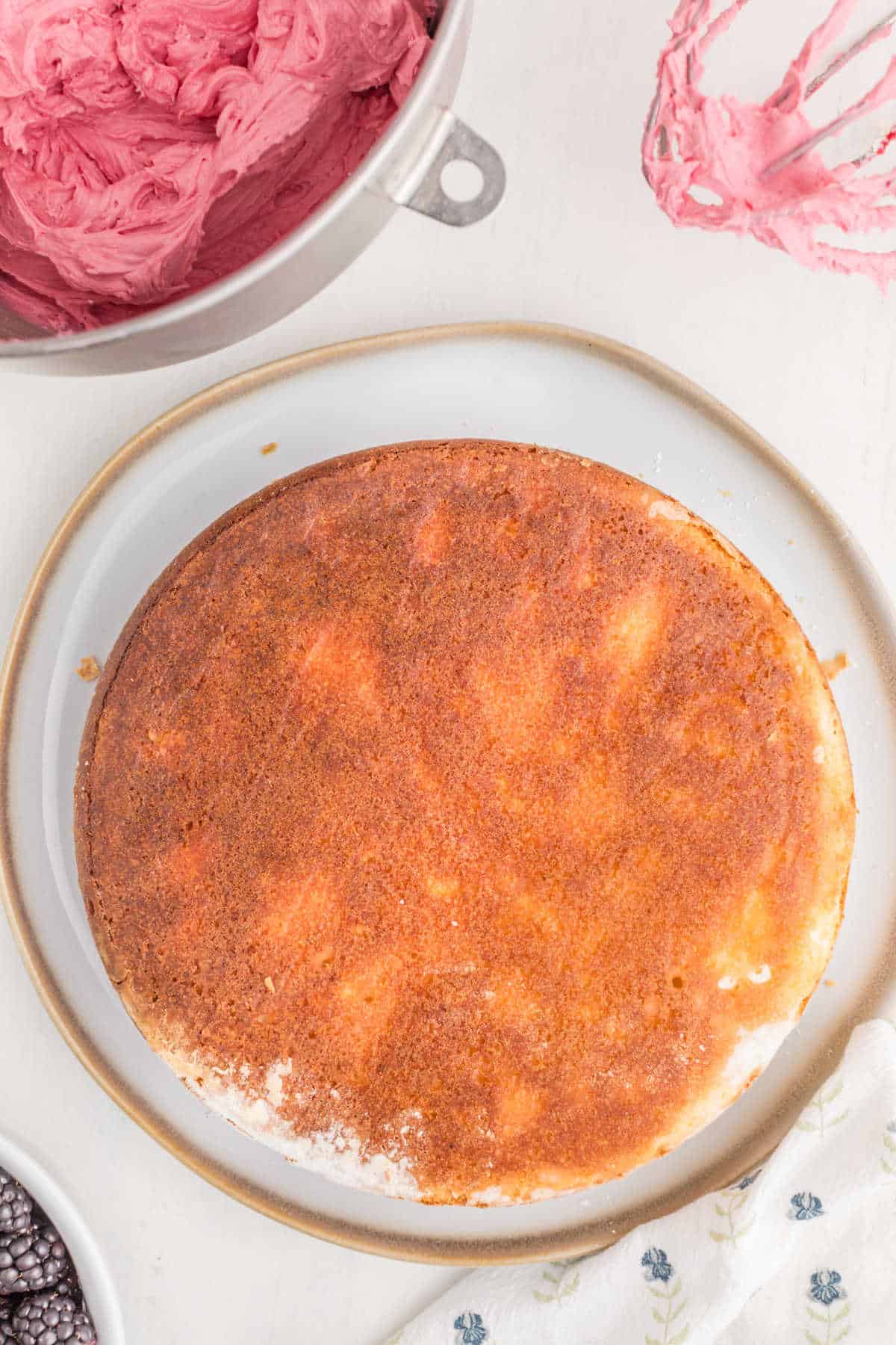 A plain round cake sits on a plate, next to a bowl of pink frosting and an electric mixer whisk coated with frosting.
