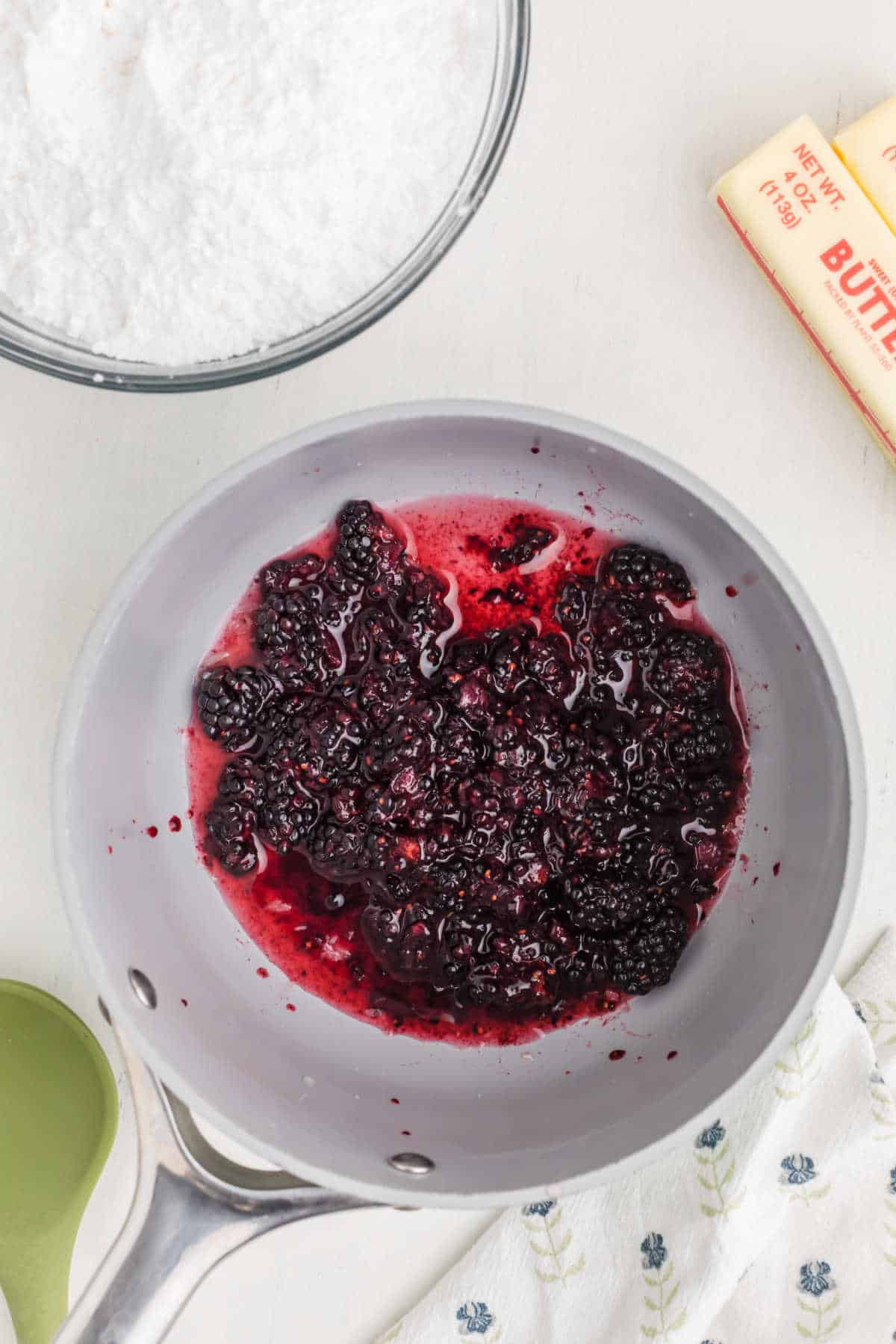 A saucepan with blackberries and juice, next to a bowl of powdered sugar and a stick of butter on a white surface.