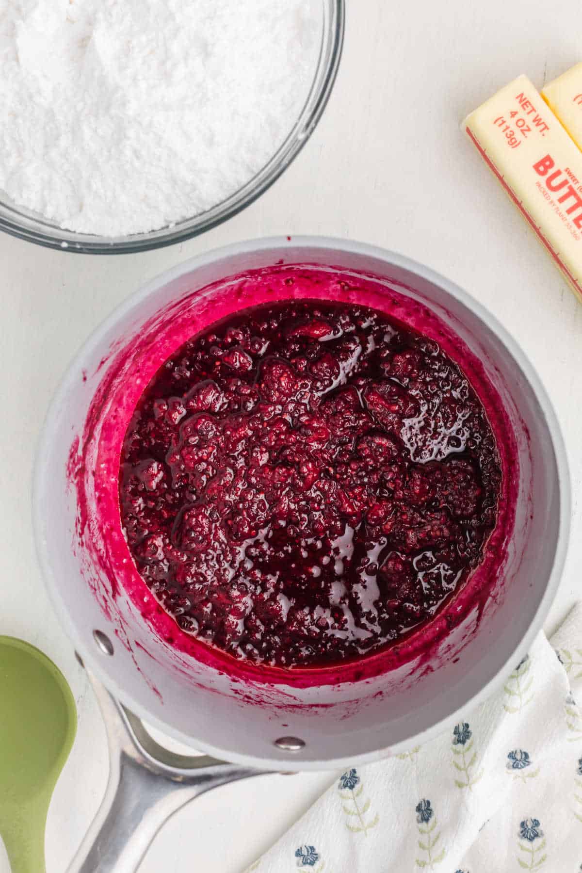 A saucepan filled with a thick, dark berry mixture sits on a white surface next to a bowl of powdered sugar, a stick of butter, and a green spatula.