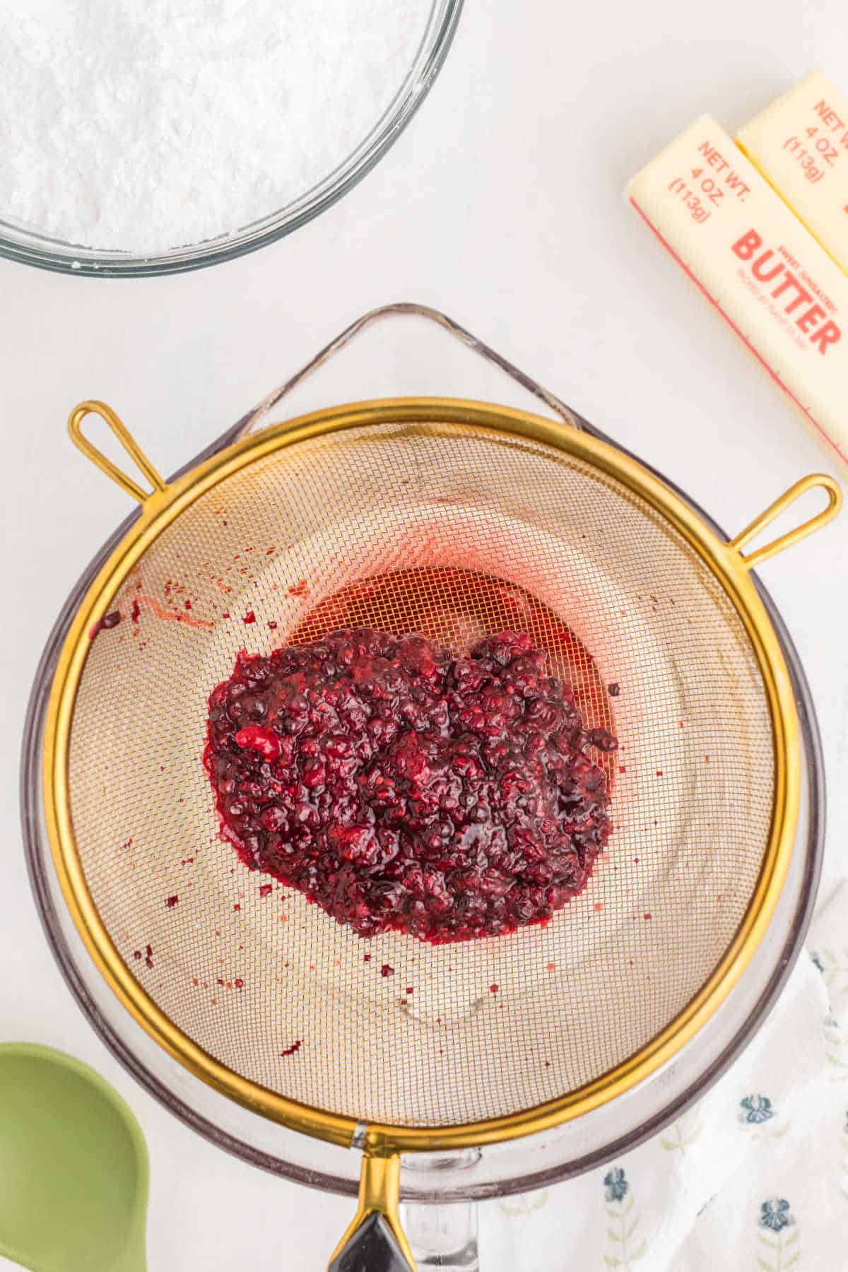 A metal strainer holds mashed berries over a glass bowl, with butter sticks, a bowl of sugar, and a green measuring cup nearby on a white surface.