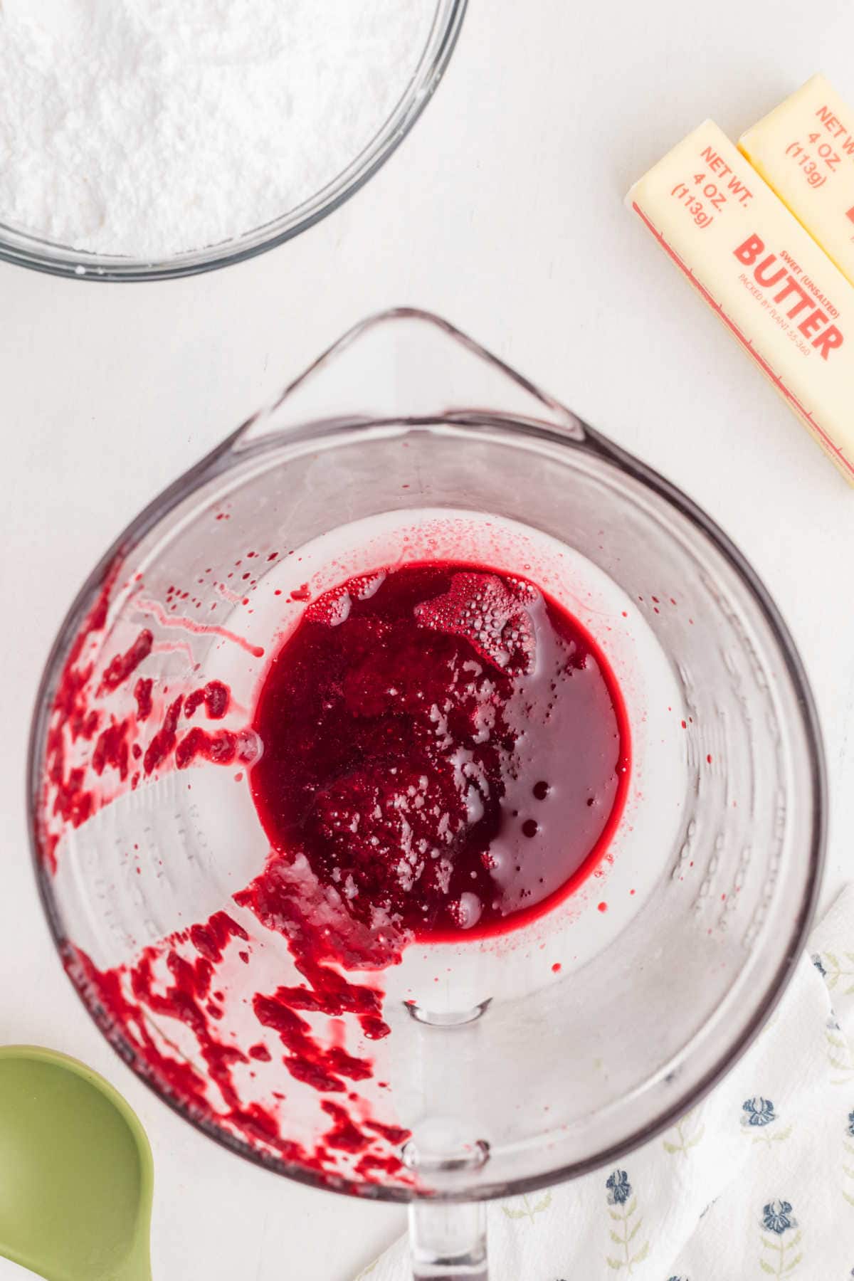 A glass mixing bowl with red liquid and residue, surrounded by butter sticks, a green measuring cup, and a bowl of powdered sugar on a white surface.