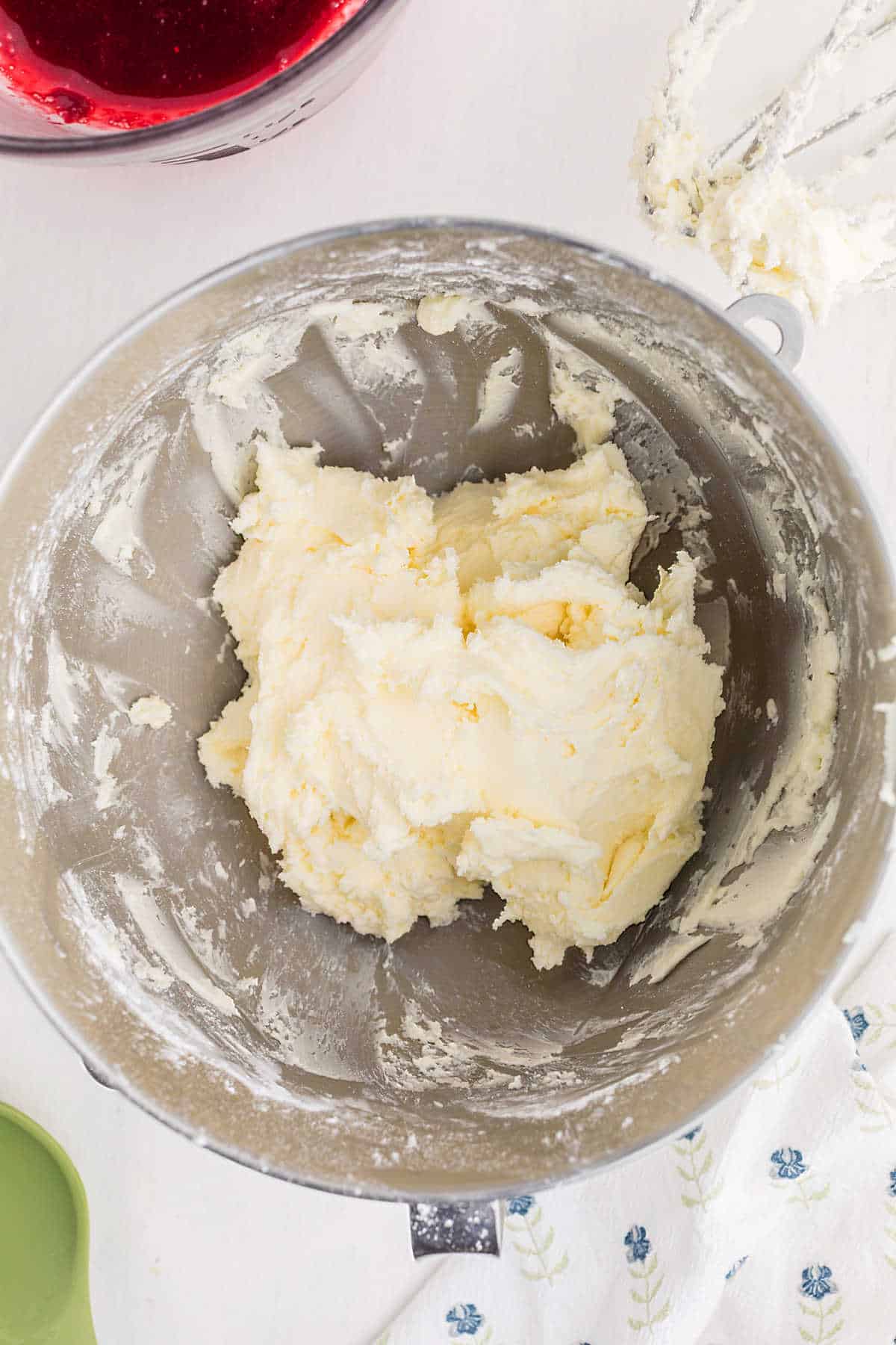 A metal mixing bowl contains creamed butter and sugar, with a stand mixer attachment resting nearby. A bowl with a red mixture is partially visible at the top.