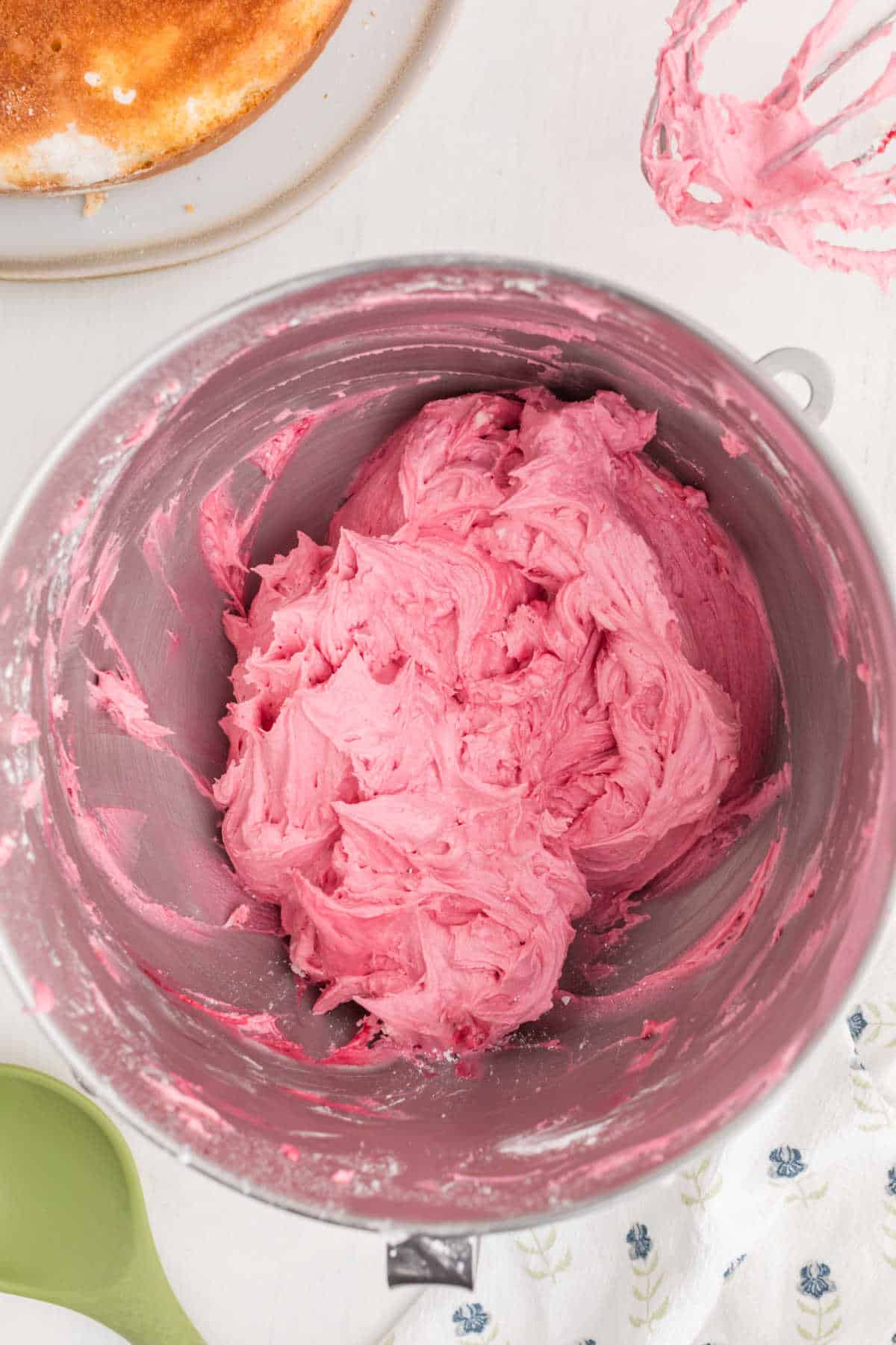 A mixing bowl filled with whipped pink frosting sits on a white surface, with a green spatula, a whisk coated in frosting, and a partially visible plate nearby.