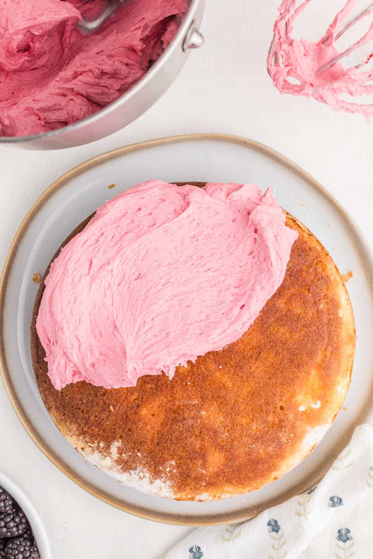 A partially frosted round cake with a large dollop of pink icing on top, surrounded by a bowl of icing, a whisk, and a white napkin with a floral pattern.