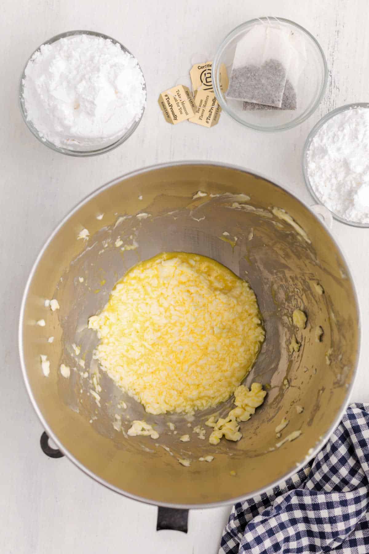 A mixing bowl with creamed butter and sugar, surrounded by bowls of powdered sugar, tea bags, and packets—essentials for making Chai Shortbread Cookies—on a light surface with a checkered cloth.