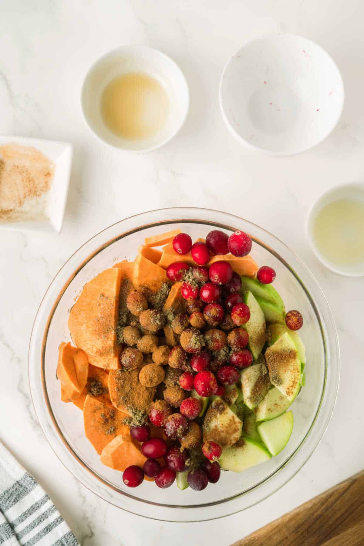 A glass bowl contains sliced sweet potatoes, green apples, and cranberries topped with brown sugar and spices; small bowls with liquids and a plate with cinnamon are nearby.