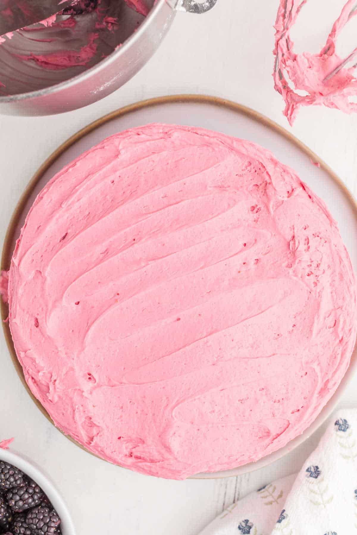 A round cake covered with smooth pink frosting sits on a plate, surrounded by baking tools and a bowl of blackberries.