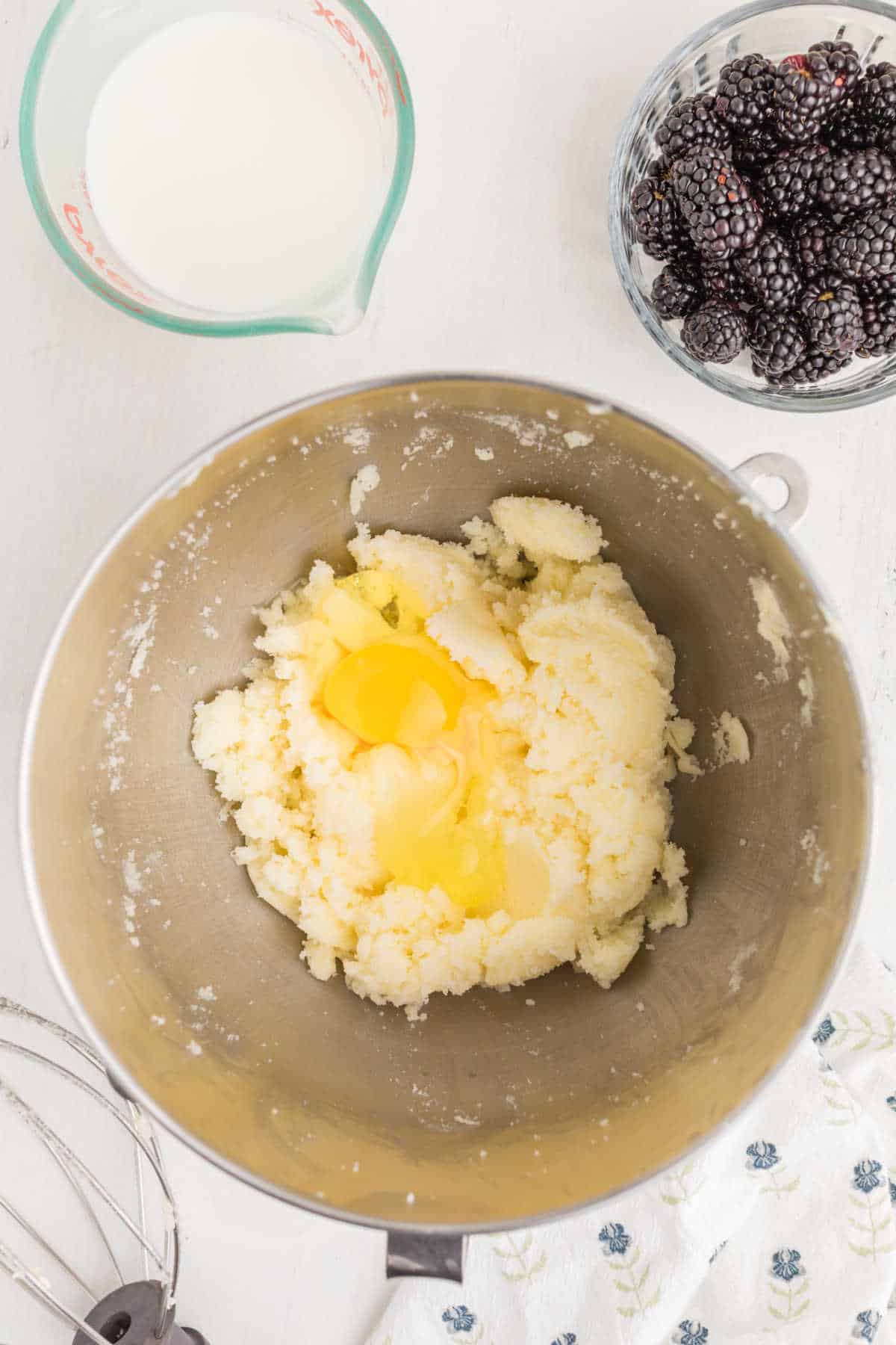 A mixing bowl with softened butter, sugar, and eggs sits on a counter beside a bowl of blackberries and a measuring cup of milk.