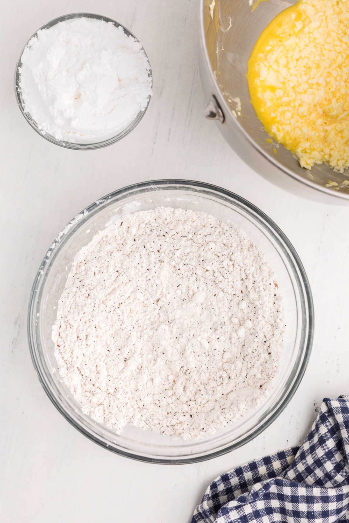 Three bowls on a white surface: one with powdered sugar, one with a flour mixture, and one with creamed butter and sugar; a blue checkered cloth is nearby.
