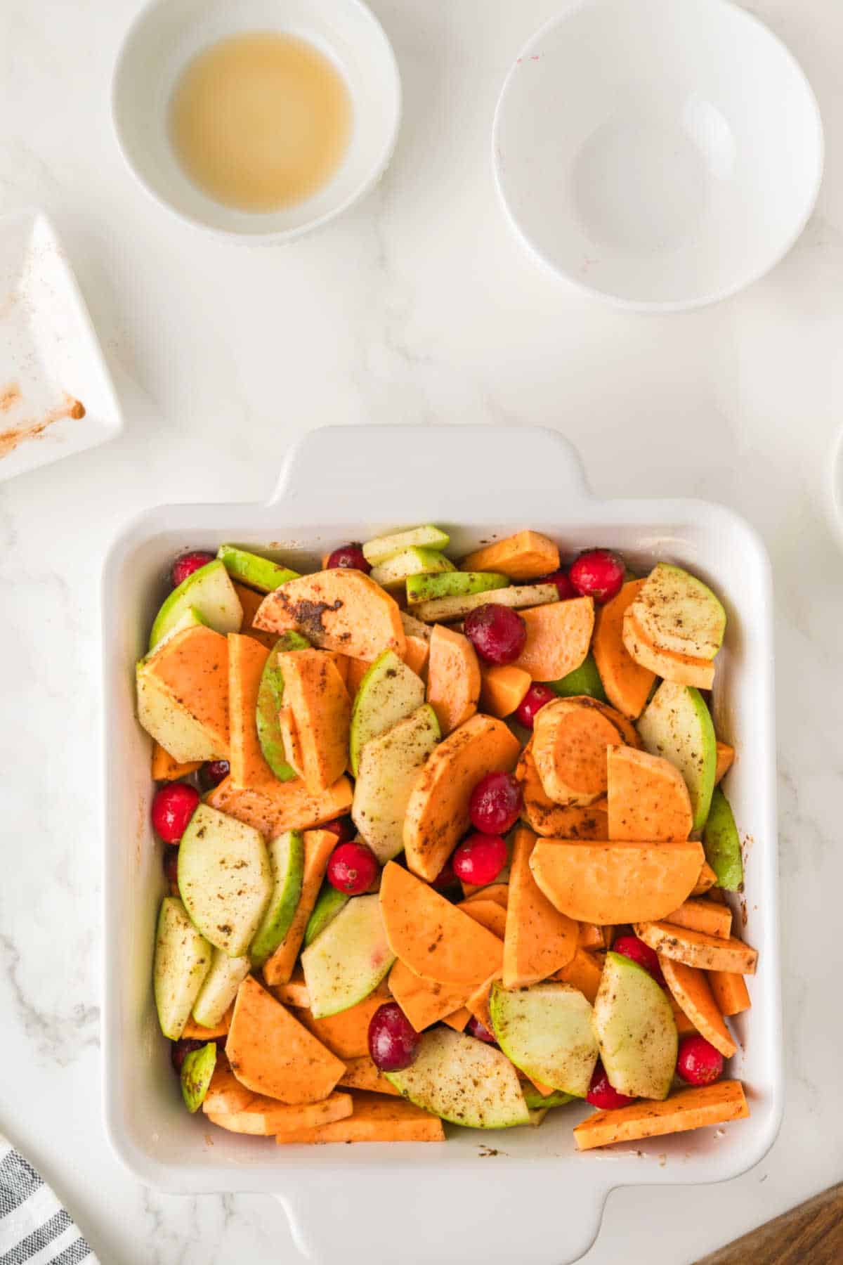 A white baking dish filled with sliced sweet potatoes, apples, and cranberries, sprinkled with spices, sits on a marble surface near empty bowls.