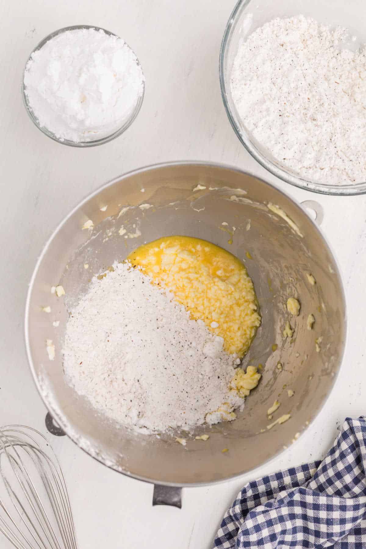 A mixing bowl with creamed butter and sugar, partly covered with flour. Nearby are bowls of flour and powdered sugar, a whisk, and a blue plaid towel on a white surface.