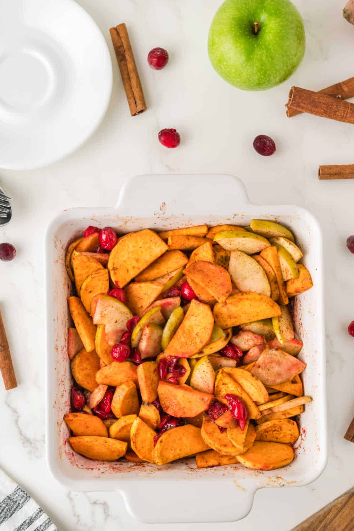 A white baking dish filled with sliced apples and cranberries, surrounded by a green apple, cinnamon sticks, and a few cranberries on a white countertop.