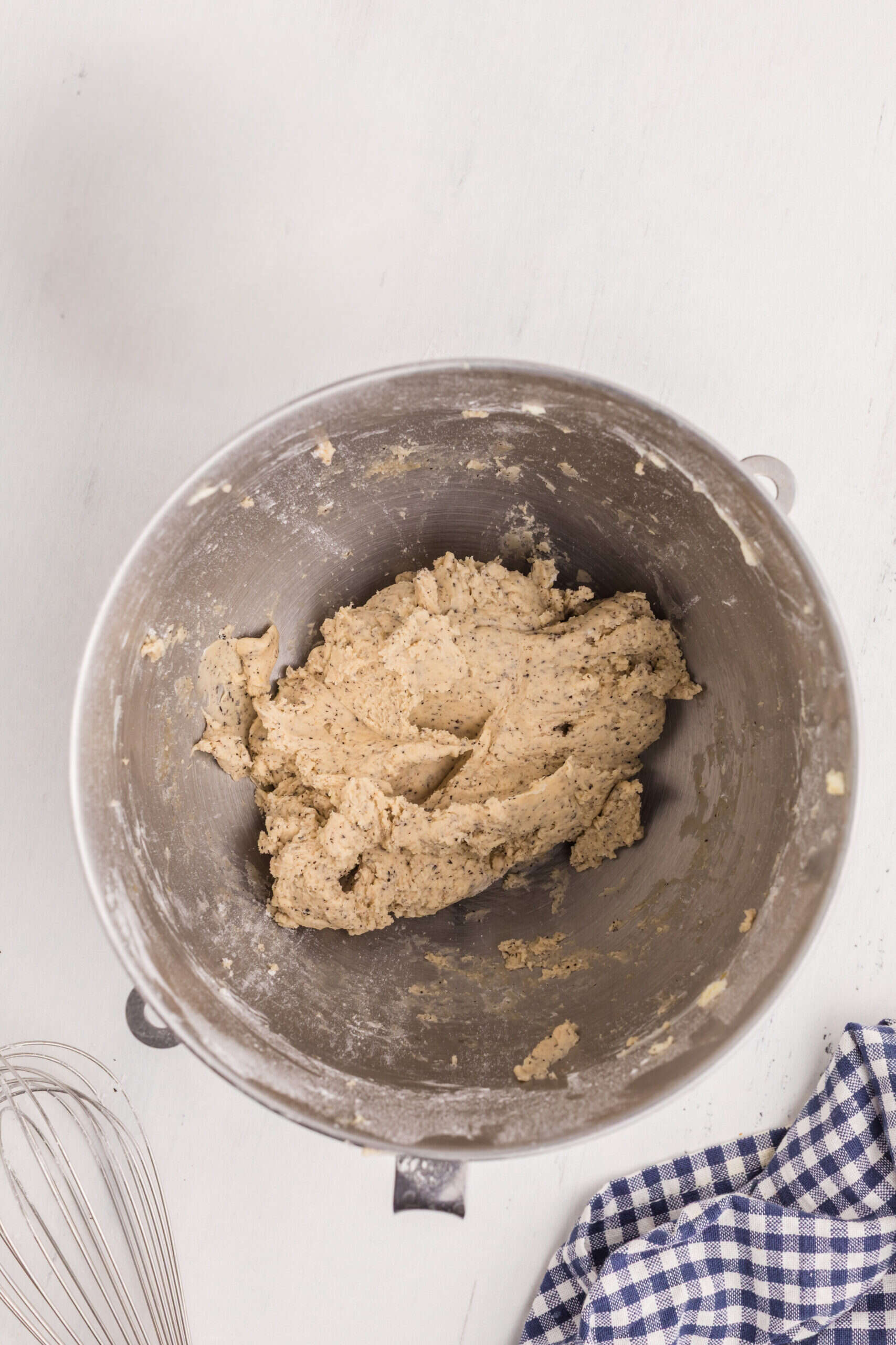 A metal mixing bowl containing thick cookie dough sits on a white surface next to a whisk and a blue and white checkered cloth.