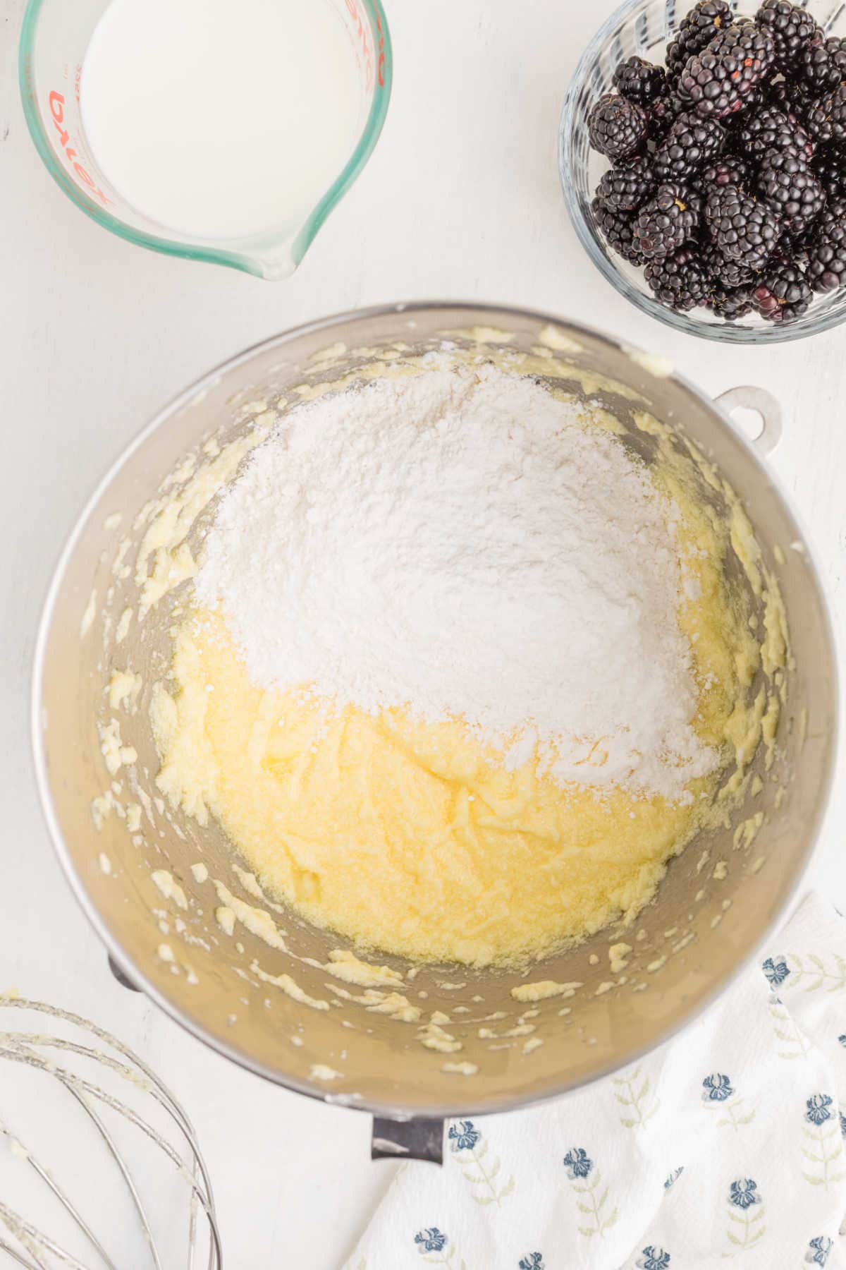 A mixing bowl with creamed butter and sugar, covered with flour, next to a bowl of blackberries and a measuring cup of milk on a white surface.
