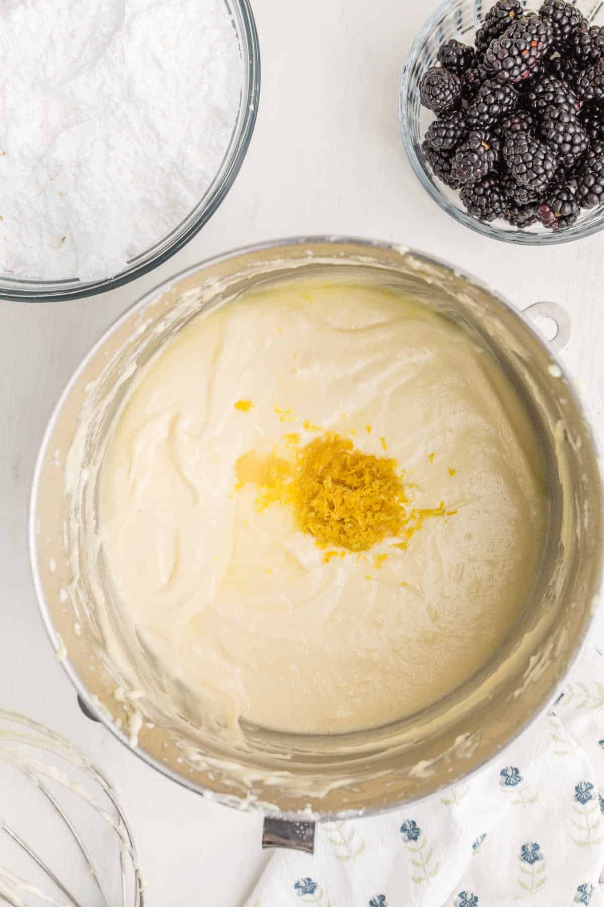 A mixing bowl with creamy batter and lemon zest on top, next to bowls of powdered sugar and blackberries on a white surface.