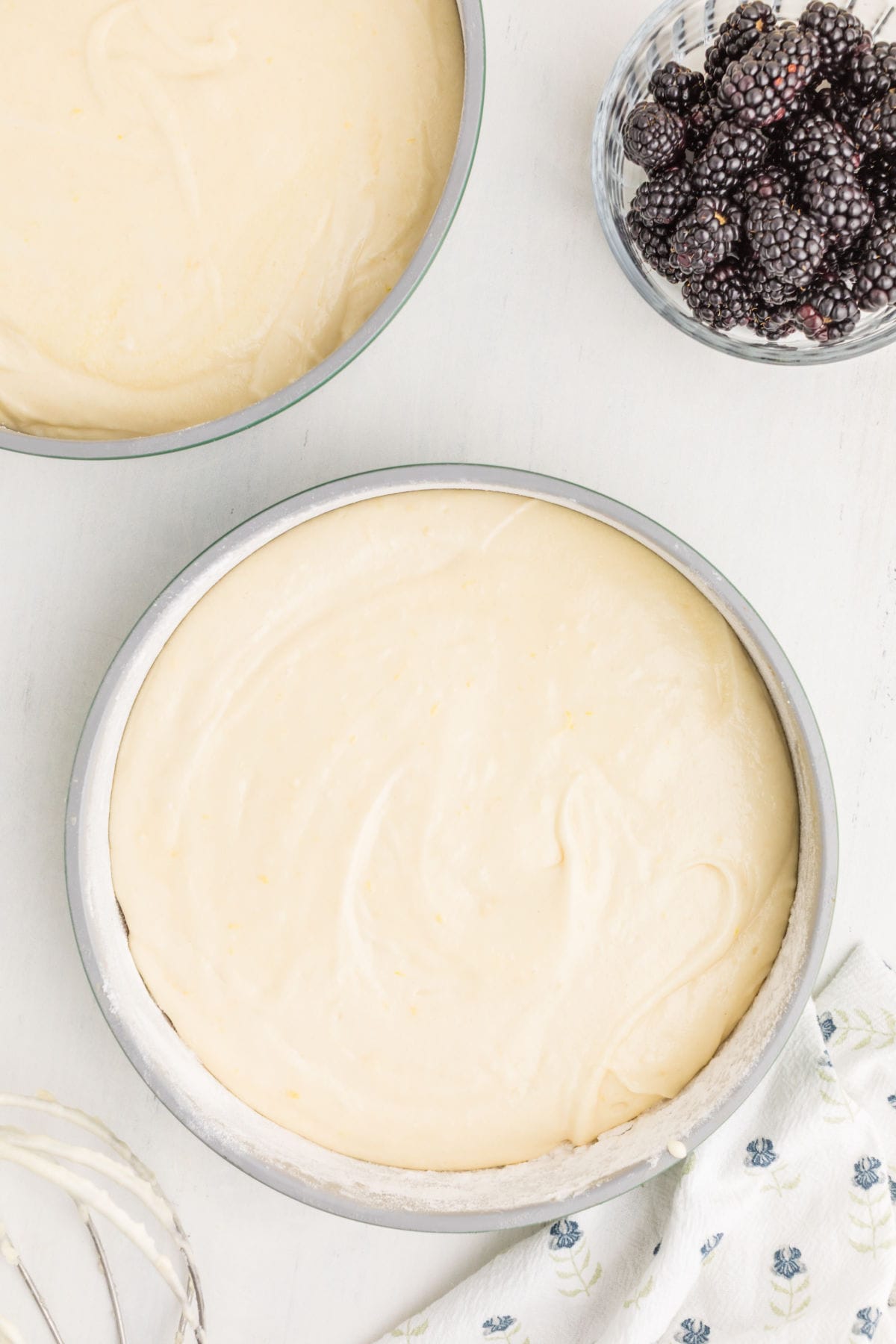 Round metal cake pans filled with cake batter, a glass bowl of blackberries, and a patterned kitchen towel on a white surface.