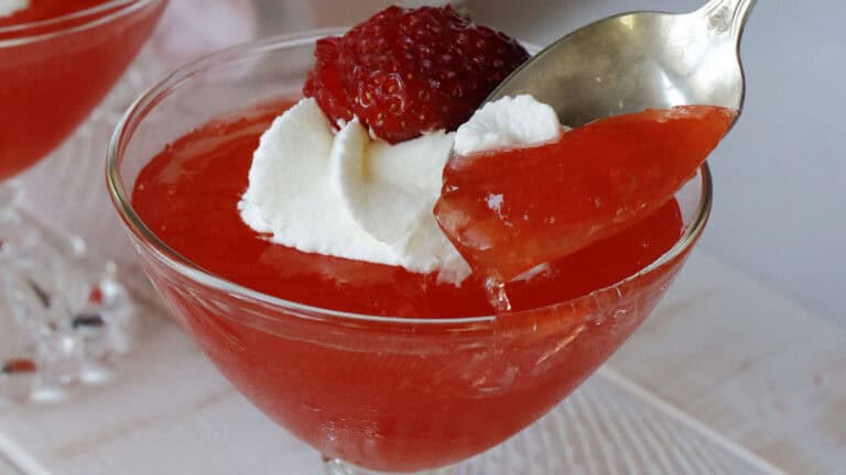 A glass dessert dish with red gelatin, topped with whipped cream and a strawberry, with a spoon lifting a portion of the gelatin.