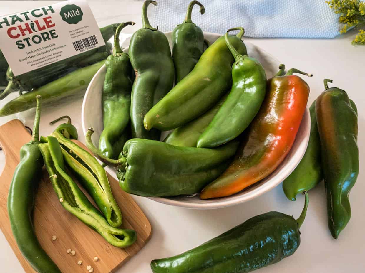 A bowl of fresh green Hatch chiles with one partially red chile, some whole and some sliced open on a cutting board, labeled as "mild.