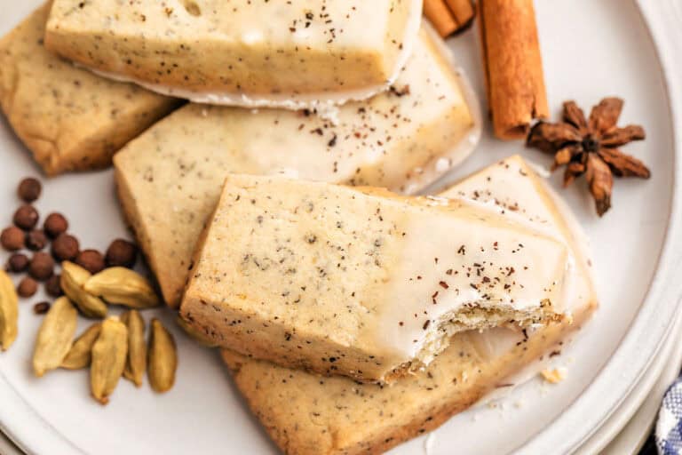 Chai shortbread cookies with chai icing, including cinnamon sticks, cardamom pods, and star anise, arranged on a white plate; one cookie has a bite taken out.
