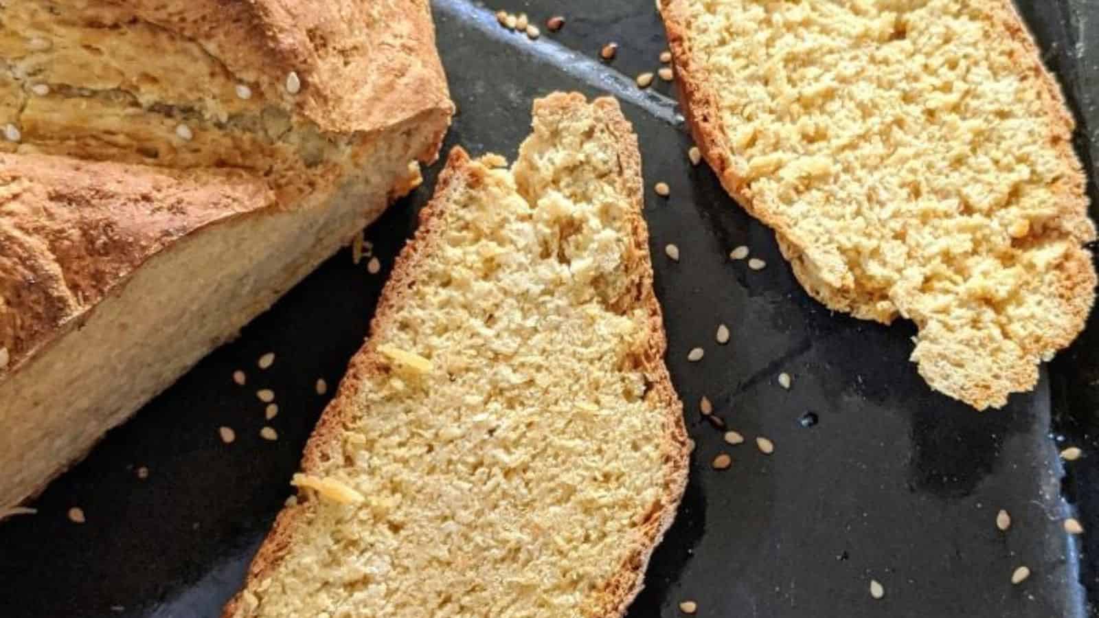 A loaf of bread and two slices are displayed on a dark surface, with sesame seeds scattered around.