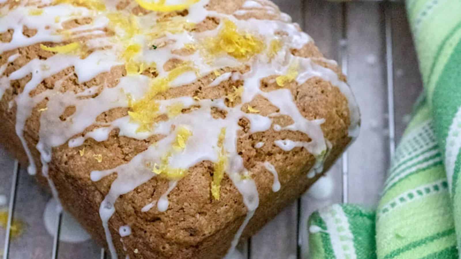 A loaf of bread topped with white icing and lemon zest sits on a cooling rack next to a green and white towel.