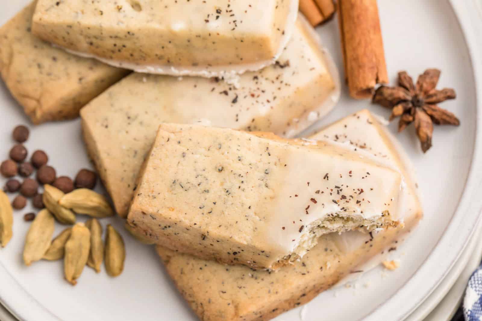 Rectangular glazed cookies with visible tea leaves, one with a bite taken out, on a plate with whole spices including cardamom, allspice, cinnamon, and star anise.