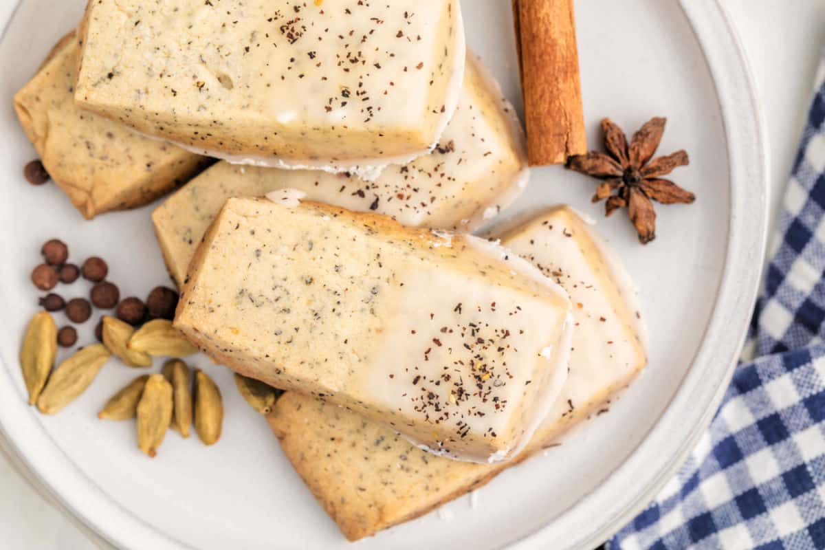 Rectangular iced cookies sprinkled with tea leaves on a white plate, surrounded by cardamom pods, allspice, star anise, and a cinnamon stick. Blue plaid napkin nearby.