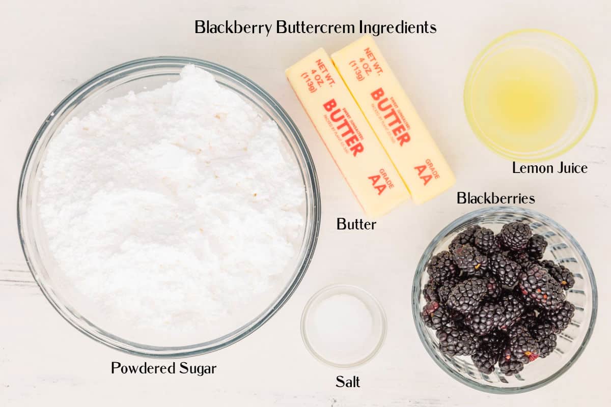 Top-down view of ingredients for blackberry buttercream: powdered sugar, butter sticks, lemon juice, blackberries, and salt, each labeled and arranged in separate bowls on a white surface.