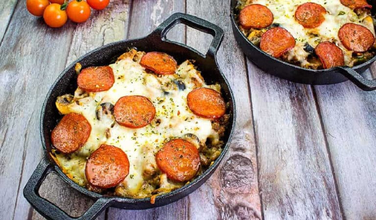 Two cast iron skillets containing baked dishes topped with melted cheese, pepperoni slices, and herbs are placed on a wooden surface. Cherry tomatoes are in the background.