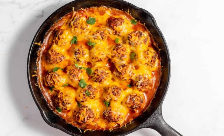 A cast iron skillet filled with baked meatballs in tomato sauce, topped with melted cheese and garnished with fresh herbs.
