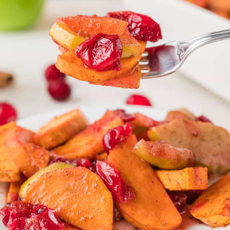 A fork holds a bite of baked sweet potatoes, apple slices, and cranberries over a plate filled with the same mixture.