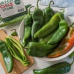 A bowl of fresh green Hatch chile peppers sits next to a cutting board with sliced chiles and a labeled bag reading "Hatch Chile Store Mild.