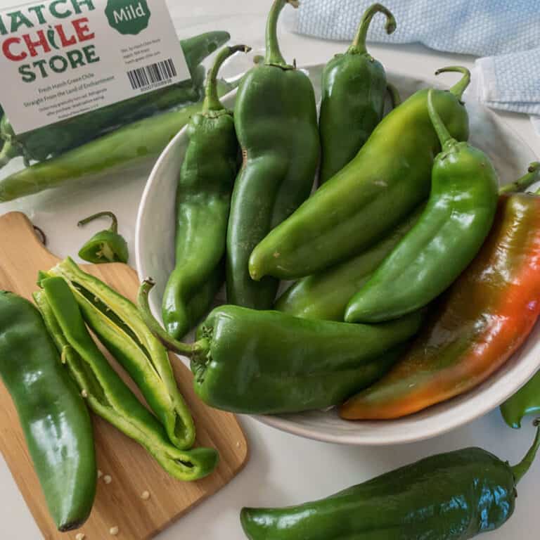 A bowl of fresh green Hatch chile peppers sits next to a cutting board with sliced chiles and a labeled bag reading "Hatch Chile Store Mild.