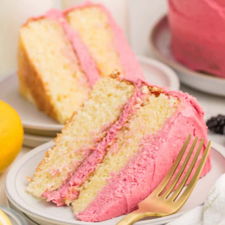A slice of yellow cake with pink frosting on a white plate, with a gold fork beside it and another cake slice in the background.