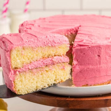 A slice of two-layer yellow cake with pink frosting is being lifted from a frosted cake on a white plate.