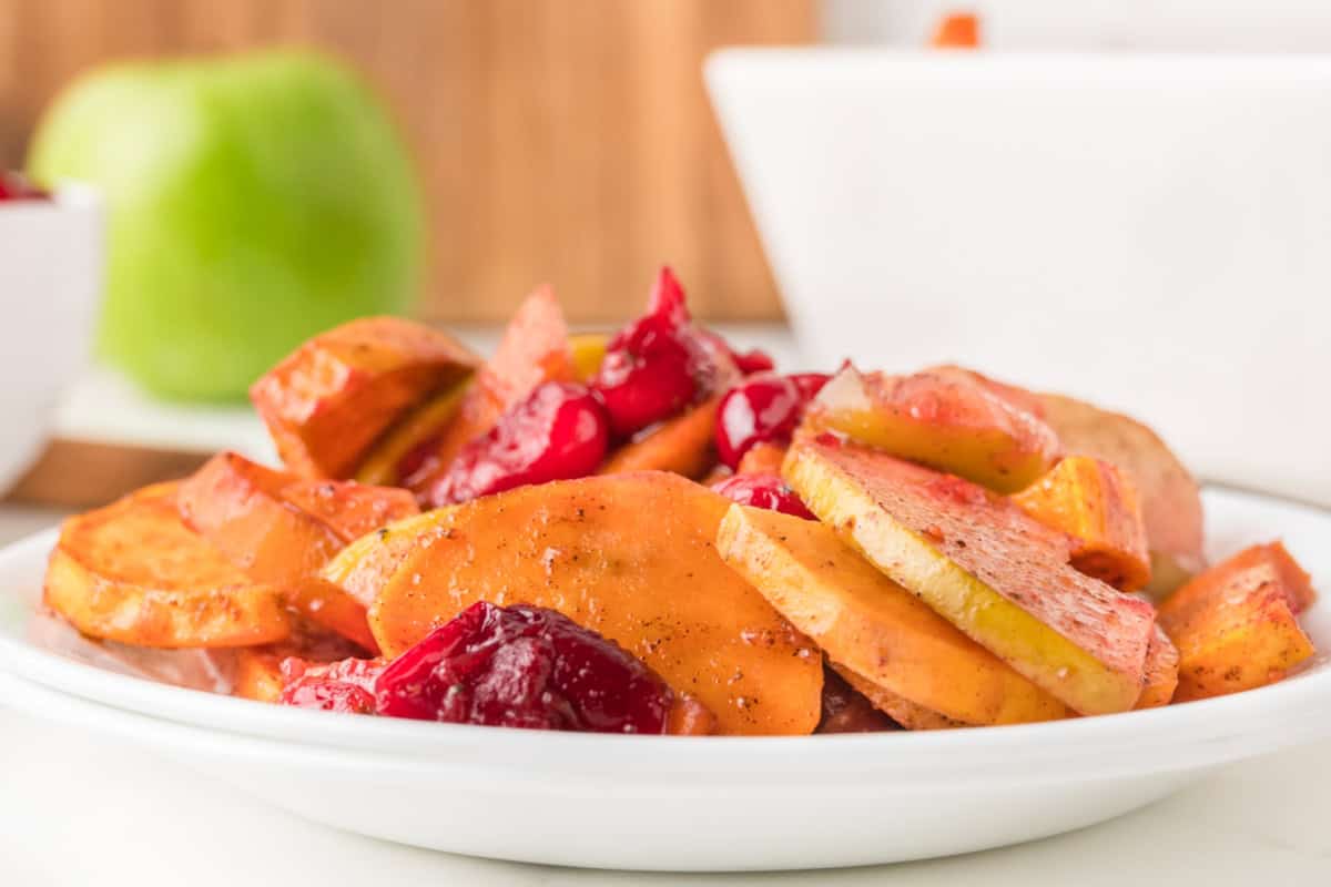 A white plate with sliced sweet potatoes and apples mixed with cranberries and spices, with a green apple and bowls in the background.