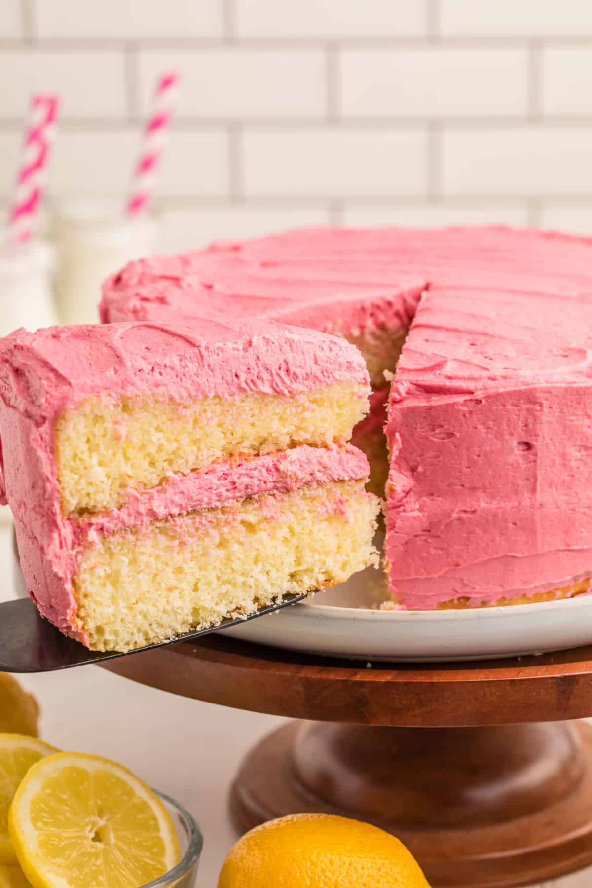 A two-layer yellow cake with pink frosting is being served on a cake stand, with lemons and a glass nearby.
