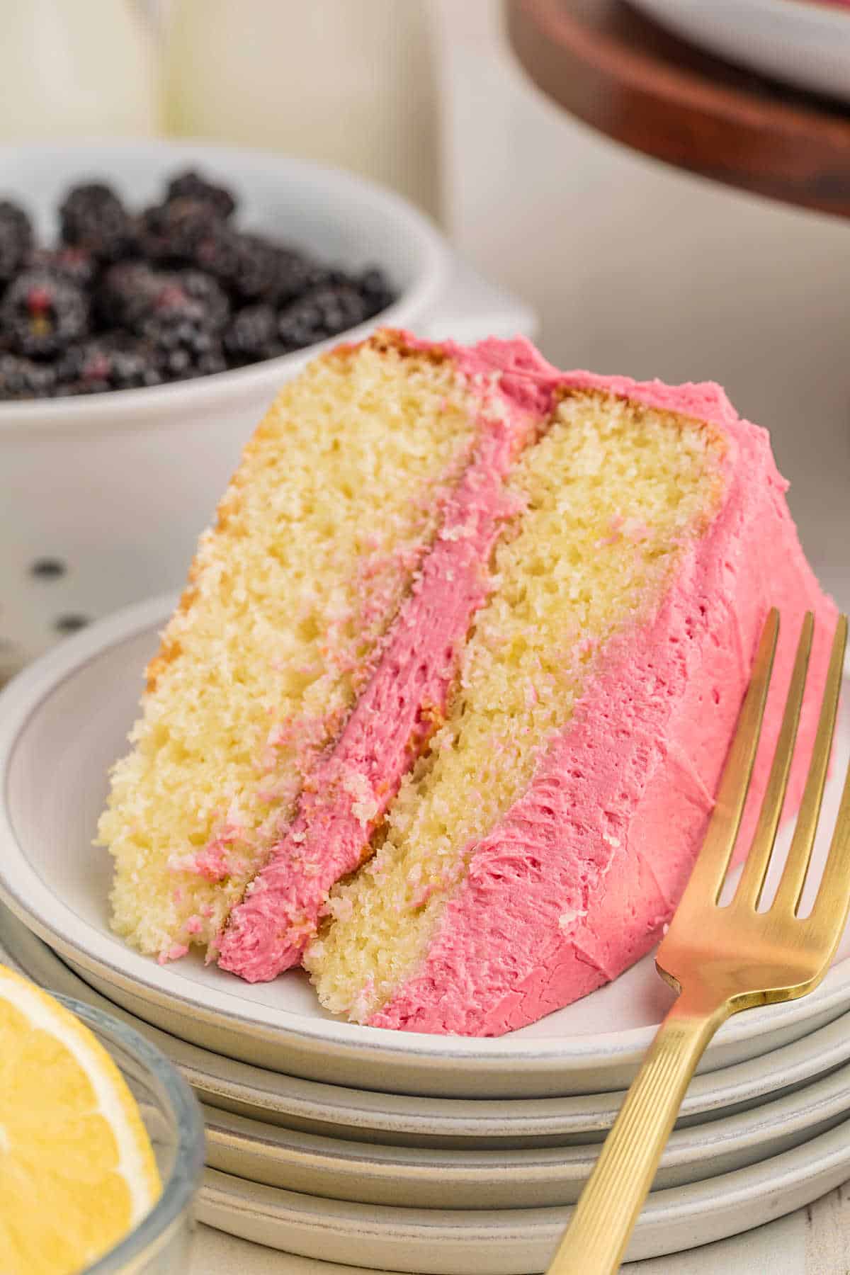 A slice of yellow layer cake with pink frosting on a white plate, next to a gold fork, with a bowl of blackberries and a lemon slice in the background.