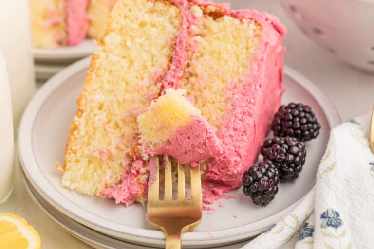 A slice of yellow cake with pink frosting and blackberries on a plate, with a fork holding a piece of cake.