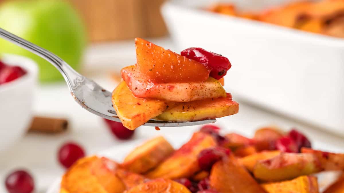 A fork holds roasted sweet potato slices and cranberries, seasoned with spices, with a bowl of fruit and a green apple blurred in the background.